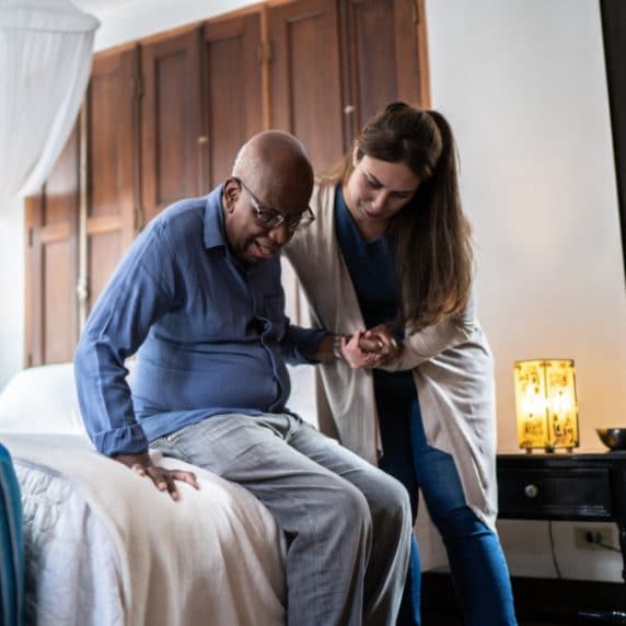 woman assisting an elderly man stand up from the bed