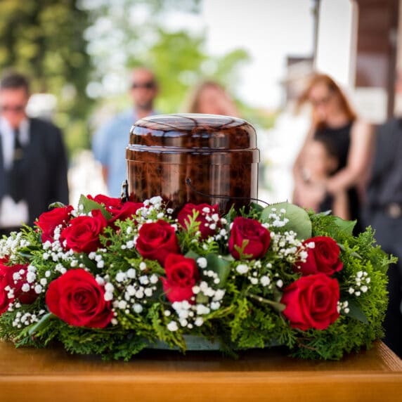 Funerary urn with ashes of dead and flowers at funeral. Burial urn decorated with flowers and people mourning in background at memorial service, sad and grieving last farewell to deceased person.