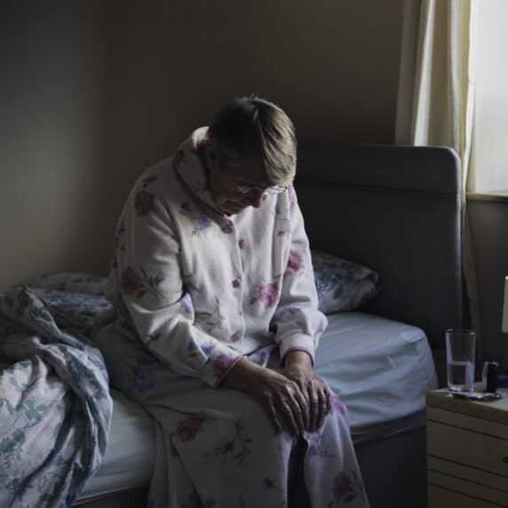 A shot of a senior woman sitting on the side of her bed with her head down looking sad. She looks like she has just woken up as is dressed in nightwear.